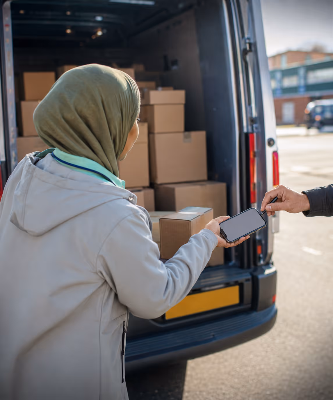 A driver capturing a customer signature on the RouteMagic app at the back of an open delivery van, parcels stacked behind.