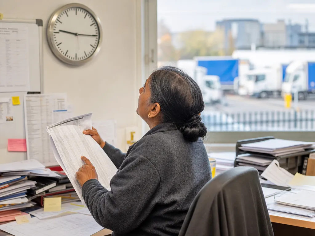 A transport desk worker squints up at a wall clock while running a finger down a long printed delivery list to guess arrival windows.