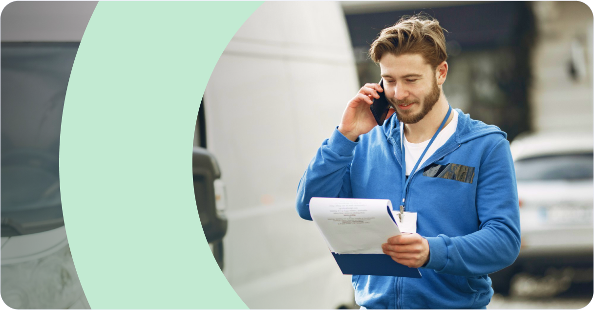 Van sales representative using smartphone while holding delivery documents with RouteMagic mobile app dashboard displaying revenue analytics & real-time tracking